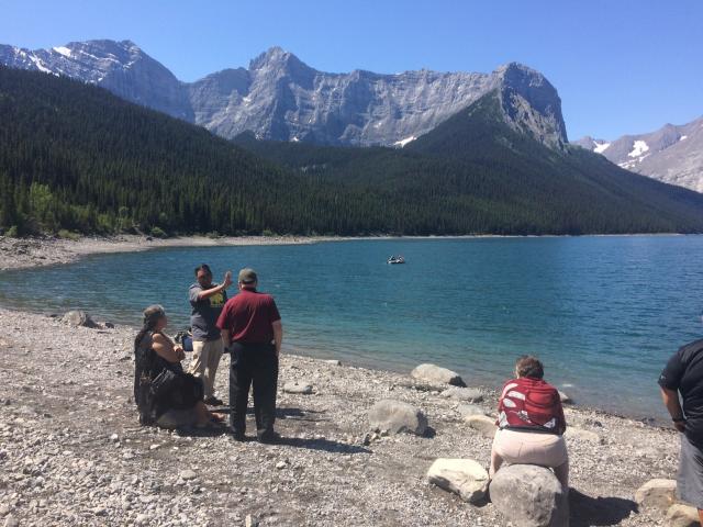Bill Snow speaking with John and Elenor Thompson (seated) at Upper Kananaskis Lake.. Bill Snow speaking with John and Elenor Thompson (seated) near a brilliant blue lake on Traditional teachings in Upper Kananaskis. Jagged grey-blue mountains overlook the scene.