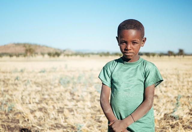 A young boy in Ethiopia, impacted by drought, helps his father tend cattle. Ethiopian Boy Holding His Arms standing in dry field
