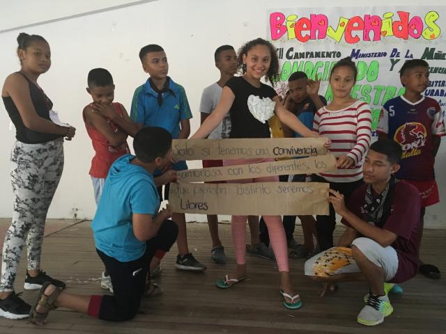 Colombian children and teenagers participate in a theatre education program. The stand in a group in front of a sign that has "Bienvenidos" (welcome).