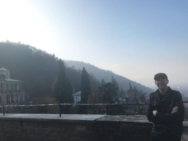 A young man who serves as a military chaplain leans against a low wall overlooking a mountainous village.