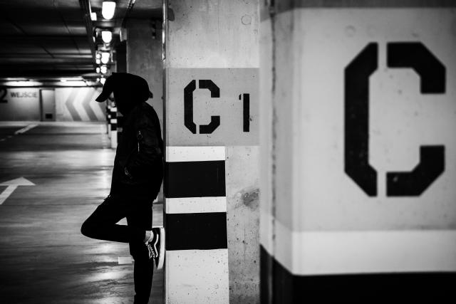 In this black and white photograph, a young man in a hoodie looks pensive while he leans against the wall in a subway station. 