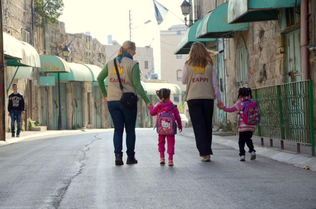 Two adults in EAPPI vests accompany two children walking down the sidewalk
