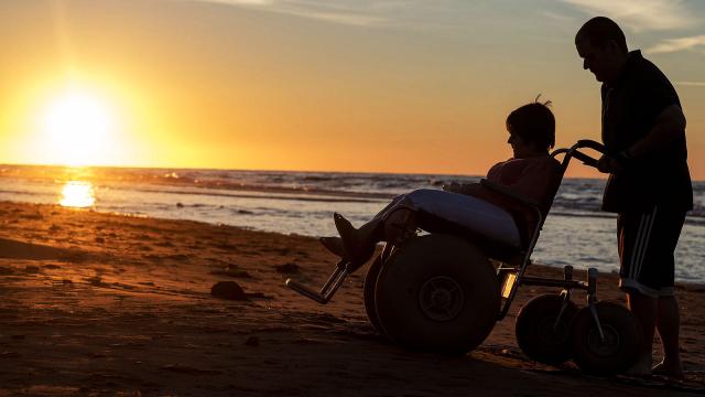 A person in a beach wheelchair crosses the sand while silhouetted by the brillant sunset.