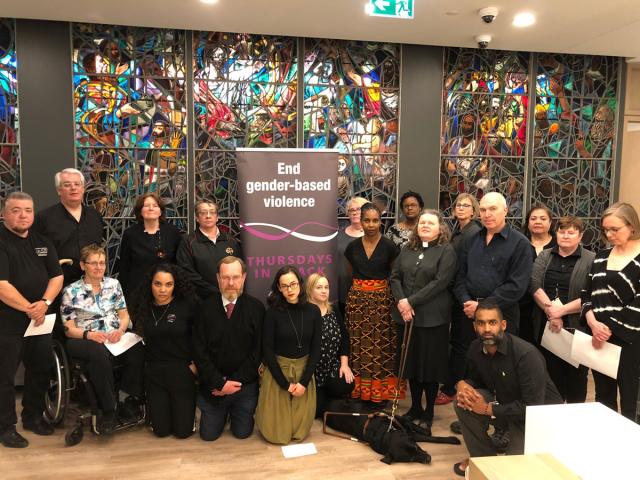This photo shows a group of 19 diverse people of differing backgrounds and types of abilities in front of a large multi-coloured stained glass. Included in the group are those who participated in the conversation on inclusion. conversation