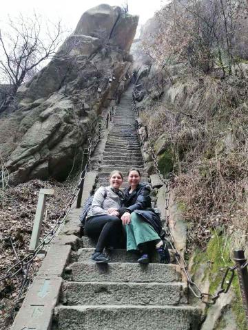 Two young women sit together on the stone staircase of a huge mountain in China.