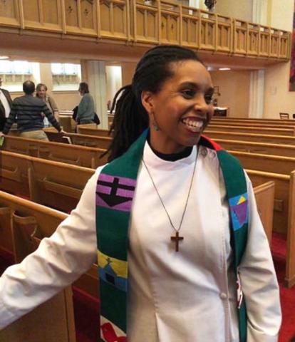 Rev. B. Maya Douglas, a Black woman, stands smiling among the pews of her church, wearing an alb and colourful embroidered stole.