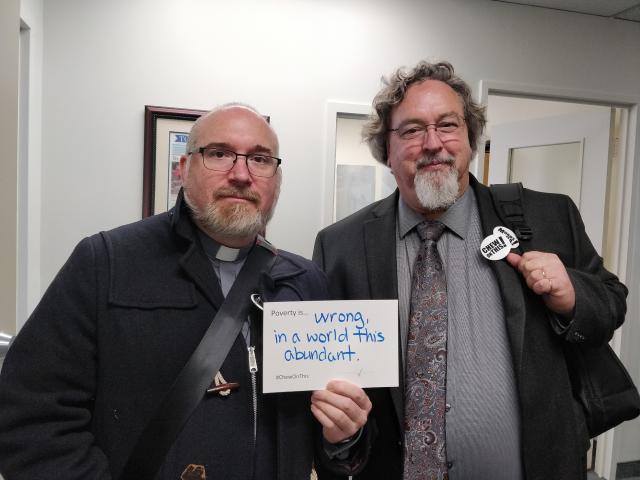 Moderator Richard Bott holds a sign that says "poverty is wrong, in a world this abundant" and stands to the left of a colleague from the Canadian Council of Churches.