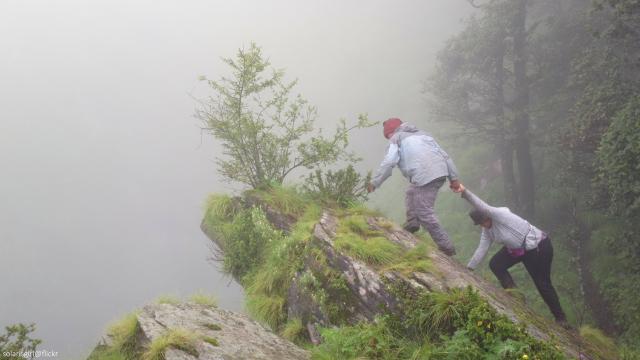 Two people work collaboratively to climb over a rock on a foggy forest mountain.