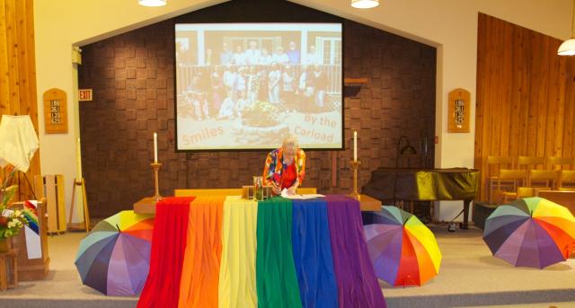A minister stands in front of a church altar decorated for a Pride celebration with rainbows.