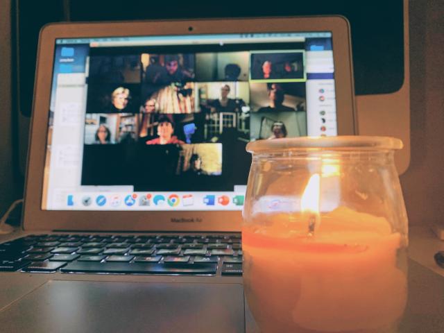 A lit candle is in front of a laptop that displays a Zoom meeting in progress.