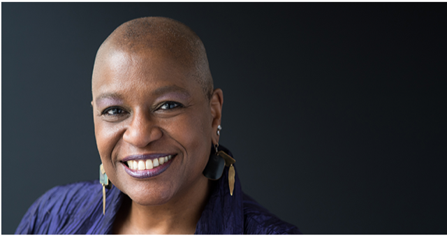 Dr. Velda Love of the United Church of Christ, a Black woman in an indigo wrap, with very closely cropped hair, a brilliant smile, and long earrings, looks directly at the camera.