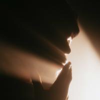 A silhouette of a person touching their lips in prayer, as light pours between their fingers.