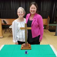 Rev. Leigh Sinclair (left) and Rev. MiYeon Lim (right) smile together as they stand before a temporary altar with a wooden cross and a small candle.