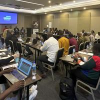 A packed conference room lined with people writing or on laptops. Four presenters sit at the front.