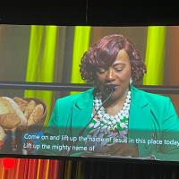 A photo of a woman speaking into a microphone projected onto a large screen