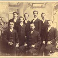 A group of men in 1892, in the formal dress of the time, pose as a group, with the front central figure holding an engraved wooden staff.