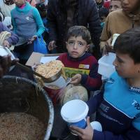 A group of Palestinian children await food, as it is being scooped out of a large pot.