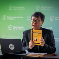 A man in mid-speech holds up a book, sitting down before a laptop with a United Church sticker on it at a desk.