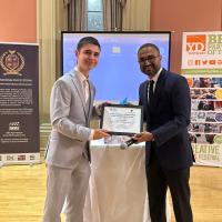 A young man in a suit receives a plaque from another man in a suit at an awards ceremony.