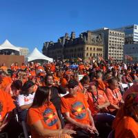 A large crowd of people wearing orange shirts sitting outside of the Canadian Parliament buildings.