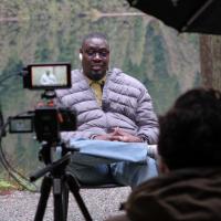 A man being filmed talking on a dock in front of a lake