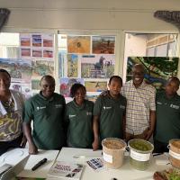 A group of individuals smiling in front of a table holding produce and grains, with photos of farming in action behind them