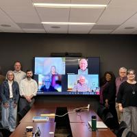 Nine people pose in a boardroom around a tv screen featuring more smiling in an online meeting