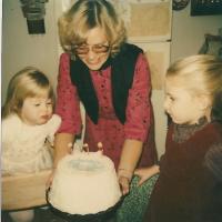 Two young girls celebrate a birthday with cake held by an adult woman in the 1980s