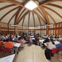 Several dozen people gather at desks in a large room with vaulted timber ceilings.