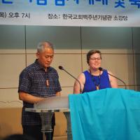 A Korean person and white person speak at a podium with a banner in Korean behind them