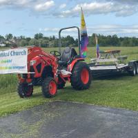 A rainbow picnic table is carried on a trailer by a tractor bearing a "Sackville United Church" banner